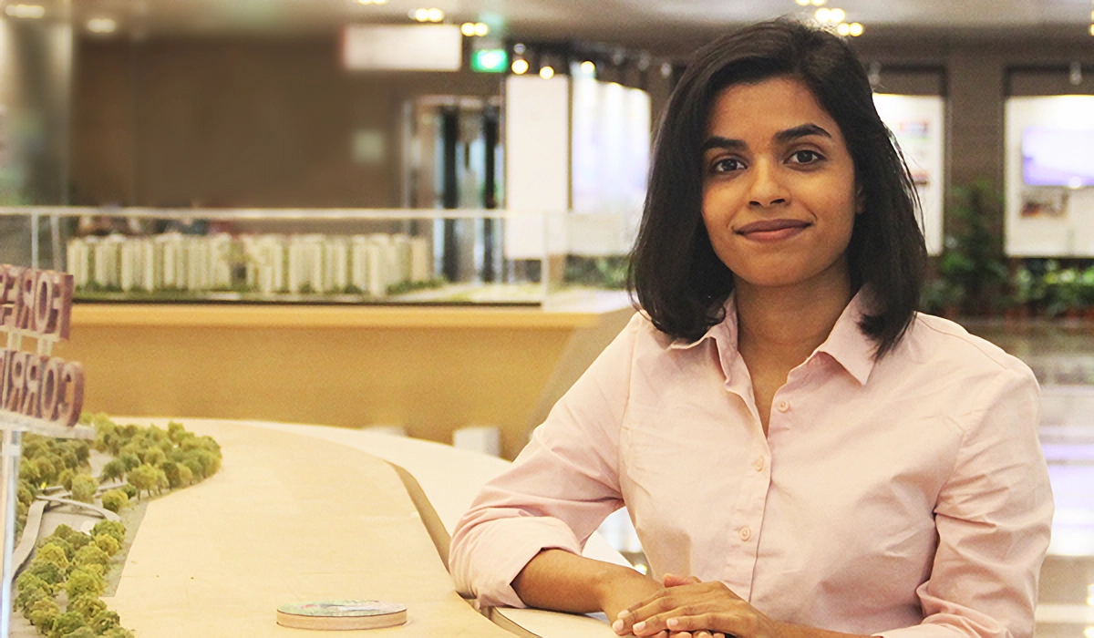 A young woman in a pink shirt sits beside a detailed architectural scale model of a city with buildings and trees.